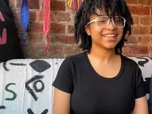 A smiling, light skinned, young Black woman, Zen Spencer, seen from the waist up wearing a short-sleeved black leotard and white aviator glasses. Her hair is styled in 2-strand twists and she’s sitting on a coach covered by white material with black abstract markings. Behind her head is a brick wall with colorful fabric art pieces strung about.
